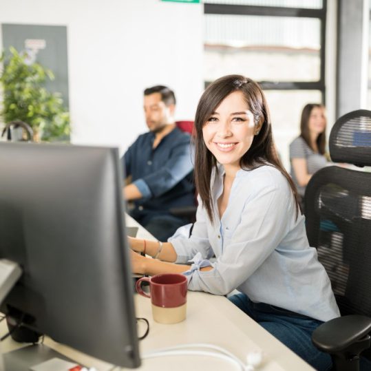 retrato-joven-latina-sonriente-sentada-su-escritorio-colegas-que-trabajan-atras